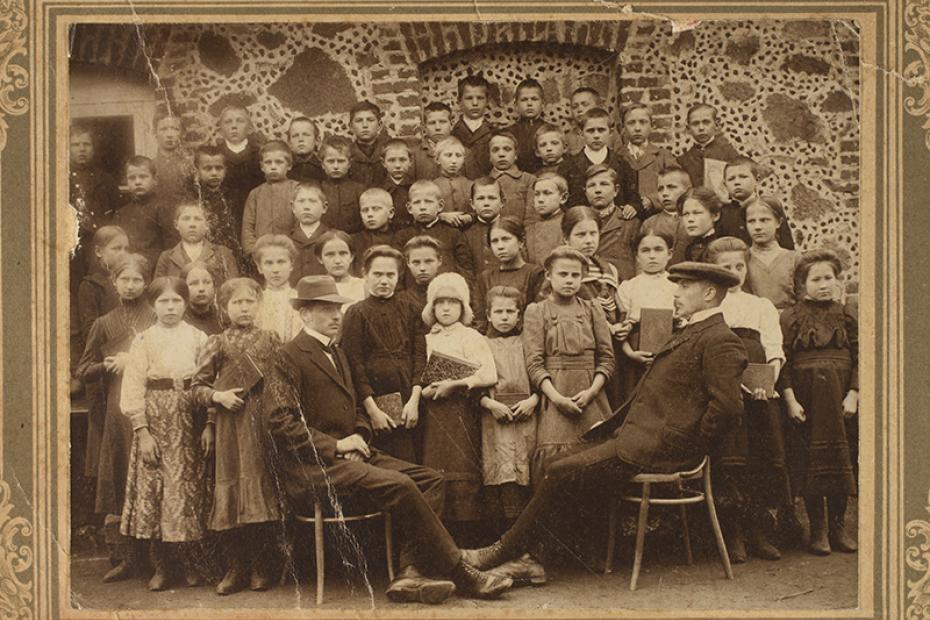Vana sepia tooniga klassipilt: suur rühm koolilapsi seisab taga, ees istuvad kaks meest ülikondades; taust on kivisein ja ajaloolise raamiga.