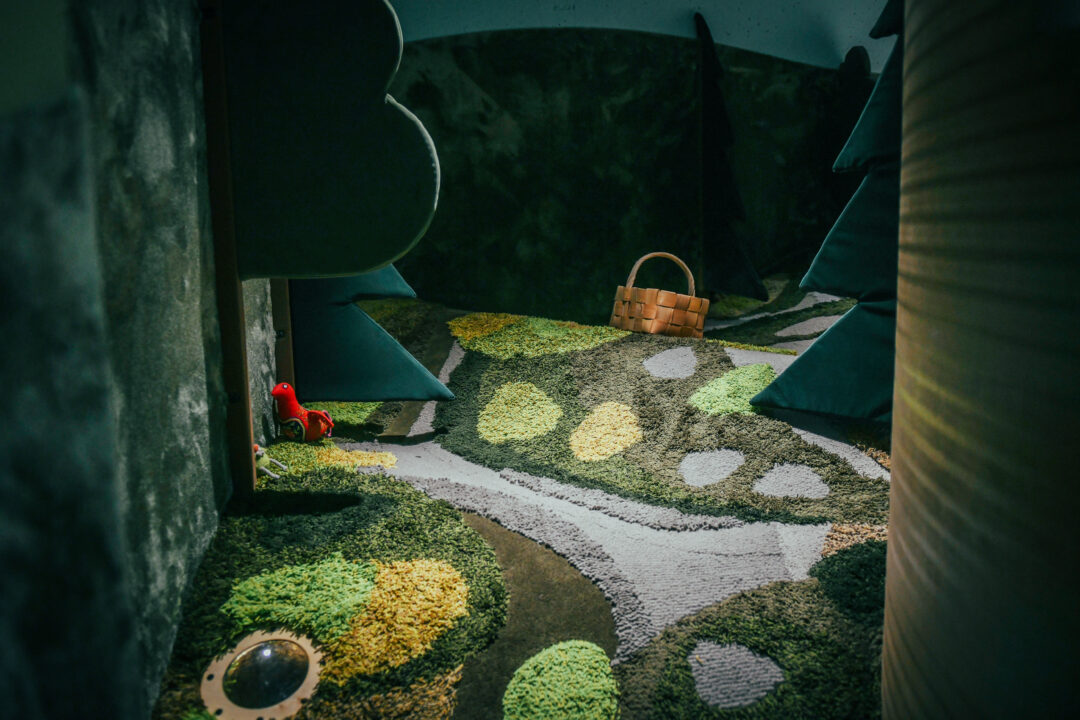 Hämara valgusega ruum, mille põrandal on metsamaastikku meenutav rohelise, kollase ja halli toonides vaip; vasakul on väike punane mänguasi. Keskel-taga on punutud korv.